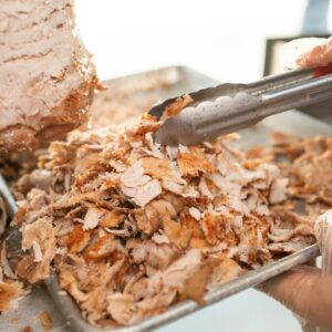 Freshly sliced shawarma meat on a tray being prepared with tongs in a kitchen setting.